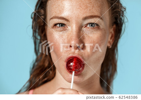Summer Freestyle. Young woman with freckles standing isolated on blue licking lollipop close-up smiling playful Summer Freestyle. Young woman with freckles standing isolated on blue licking lollipop close-up smiling playful 62548386