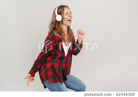 Freestyle. Young woman with freckles wearing headphones standing isolated on grey listening to music singing dancing laughing cheerful 62548389