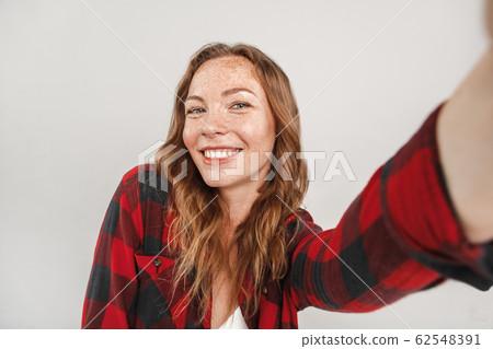 Freestyle. Young woman with freckles standing isolated on grey taking selfie on smartphone posing to camera joyful 62548391