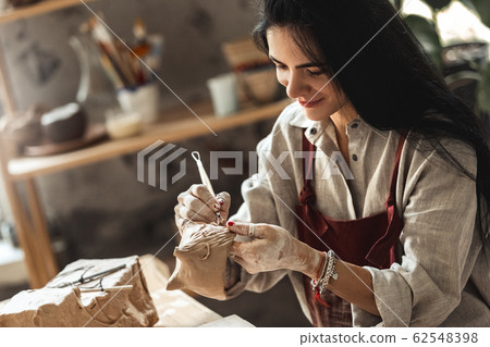 Craftsperson Concept. Young woman sitting at table making pottery indoors using modeling tool molding happy 62548398
