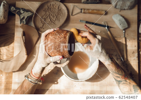 Craftsperson Concept. Young woman making pottery indoors removing dust with sponge top view close-up finishing work Craftsperson Concept. Young woman making pottery indoors removing dust with sponge top view close-up finishing work 62548407
