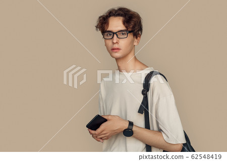 Freestyle. Man in glasses with bag standing isolated on gray holding smartphone looking camera pensive close-up Freestyle. Man in glasses with bag standing isolated on gray holding smartphone looking camera pensive close-up 62548419