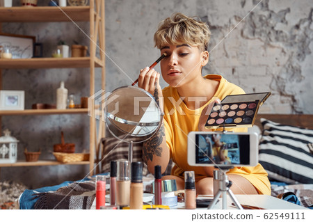 Make-up Blogger. Young woman sitting at stylish apartment recording video on smartphone looking at mirror applying eyeshadow concentrated Make-up Blogger. Young woman sitting at stylish apartment recording video on smartphone looking at mirror applying eyeshadow concentrated 62549111