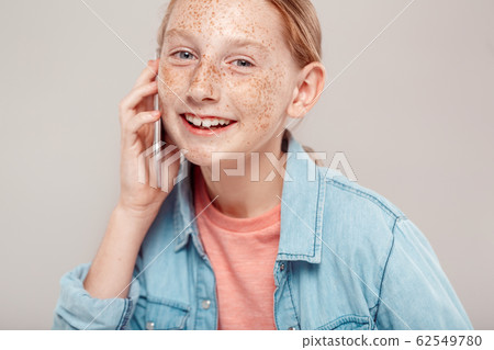 Inclusive Beauty. Girl with freckles in denim jacket standing isolated on grey talking on phone laughing joyful close-up 62549780