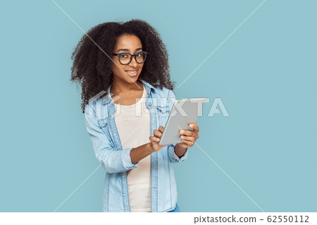 Freestyle. African girl in glasses standing isolated on gray using app on digital tablet smiling positive Freestyle. African girl in glasses standing isolated on gray using app on digital tablet smiling positive 62550112