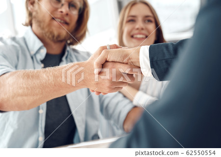 Car Salon. Young couple sitting at table man shaking hands with agent buying car close-up smiling happy Car Salon. Young couple sitting at table man shaking hands with agent buying car close-up smiling happy 62550554