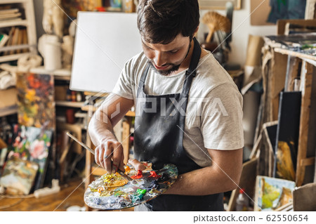 Male painter at art studio indoors mixing colors on palette with brush concentrated Male painter at art studio indoors mixing colors on palette with brush concentrated 62550654
