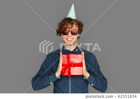 Celebration Concept. Teenager boy in birthday cap and sunglasses standing isolated on grey showing present to camera smiling toothy Celebration Concept. Teenager boy in birthday cap and sunglasses standing isolated on grey showing present to camera smiling toothy 62551834