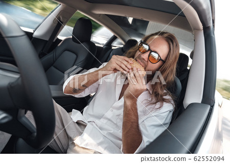 Mode of Transport. Bearded man with long hair in sunglasses traveling sitting inside electric car driving on autopilot eating hamburger joyful 62552094