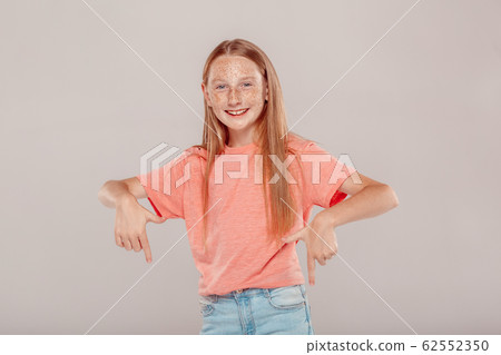 Inclusive Beauty. Girl with freckles standing isolated on grey pointing down at copy space smiling cheerful Inclusive Beauty. Girl with freckles standing isolated on grey pointing down at copy space smiling cheerful 62552350