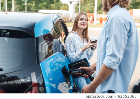Traveling. Young couple traveling by electric car stopping at charging station boyfriend plugging in cable talking with girlfriend drinking hot coffee joyful Traveling. Young couple traveling by electric car stopping at charging station boyfriend plugging in cable talking with girlfriend drinking hot coffee joyful 62552363