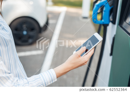 Transportation. Young woman on electric car having stop at charging station browsing smartphone while getting car fully charged Transportation. Young woman on electric car having stop at charging station browsing smartphone while getting car fully charged 62552364