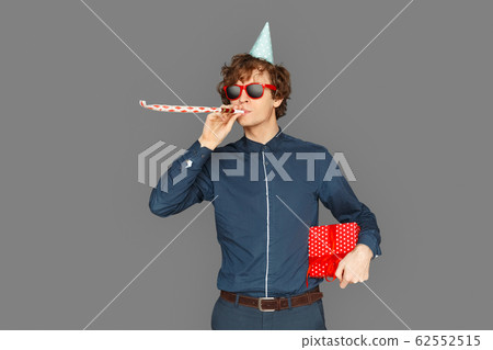 Celebration Concept. Teenager boy in birthday cap and sunglasses standing isolated on grey with present blowing party horn smiling cool Celebration Concept. Teenager boy in birthday cap and sunglasses standing isolated on grey with present blowing party horn smiling cool 62552515