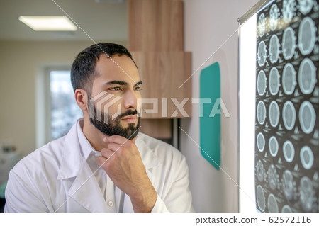 Bearded young doctor in a white robe analyzing MRI results Bearded young doctor in a white robe analyzing MRI results 62572116
