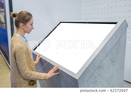 Woman using interactive empty white touchscreen display kiosk at exhibition Woman using interactive empty white touchscreen display kiosk at exhibition 62572697