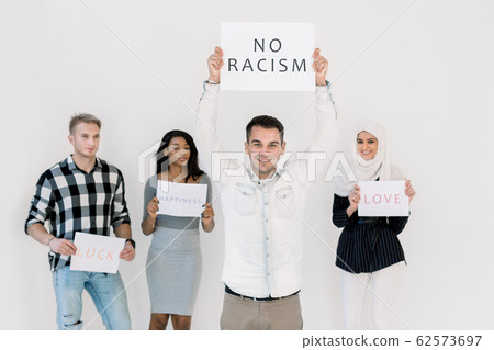 Stop racism, no racial discrimination of people. Young Caucasian man holds a poster with No racism text, while standing together with multiethnical friends with slogans on white background indoors Stop racism, no racial discrimination of people. Young Caucasian man holds a poster with No racism text, while standing together with multiethnical friends with slogans on white background indoors 62573697