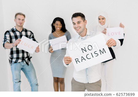 A white man in casual wear holds a poster with the No racism text in his hands, standing on white background. Three multiethnical activists chanting and holding posters with slogans on the background A white man in casual wear holds a poster with the No racism text in his hands, standing on white background. Three multiethnical activists chanting and holding posters with slogans on the background 62573699