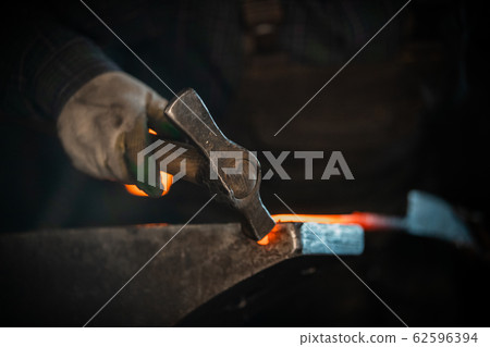 Blacksmith forging a knife out of the hot metal using a hammer - holding the knife in forceps Blacksmith forging a knife out of the hot metal using a hammer - holding the knife in forceps 62596394