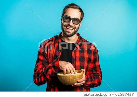 Young european manl in 3d glasses watching fascinating comedy movie, laughing and eating popcorn on blue studio background. 62597437
