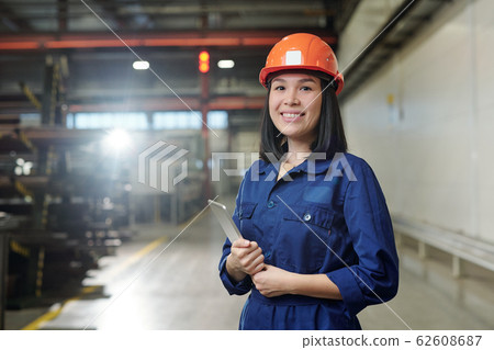 Pretty young smiling engineer in uniform and protective helmet holding tablet 62608687