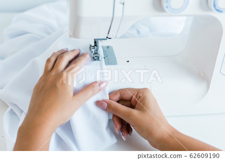 Hands of young female with white textile sitting by electric machine and sewing 62609190