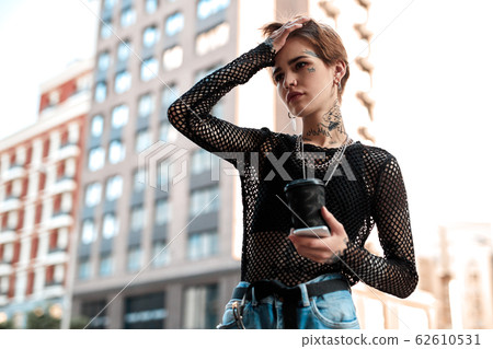 Urban Style. Young stylish woman standing outdoors with cup of coffee and phone touching head pensive 62610531