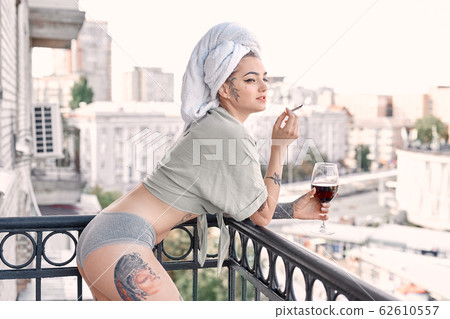 Home Leisure. Young woman in underwear and towel standing on balcony with glass of wine smoking cigarette looking aside smiling playful 62610557