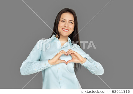 Freestyle. Young woman standing isolated on grey showing heart shape smiling cheerful Freestyle. Young woman standing isolated on grey showing heart shape smiling cheerful 62611516