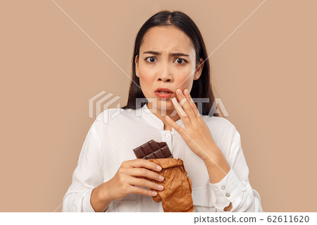 Freestyle. Young woman in shirt standing isolated on bage eating chocolate bar touching cheek having toothache Freestyle. Young woman in shirt standing isolated on bage eating chocolate bar touching cheek having toothache 62611620