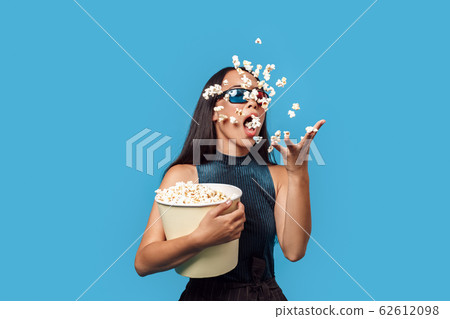 Freestyle. Young woman wearing 3d glasses standing isolated on blue eating popcorn throwing it up trick 62612098