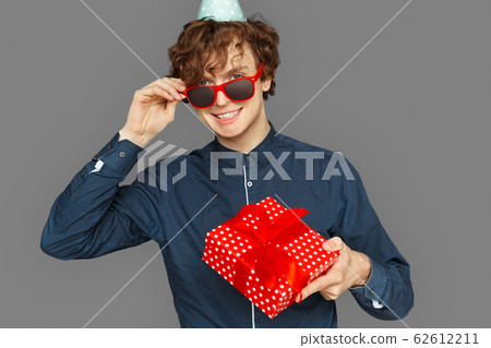 Celebration Concept. Teenager boy in birthday cap taking off sunglasses looking camera smiling playful standing isolated on grey with gift Celebration Concept. Teenager boy in birthday cap taking off sunglasses looking camera smiling playful standing isolated on grey with gift 62612211