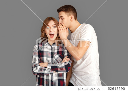 Freestyle. Man whispering secret into woman ear looking camera surprised standing isolated on grey Freestyle. Man whispering secret into woman ear looking camera surprised standing isolated on grey 62612390