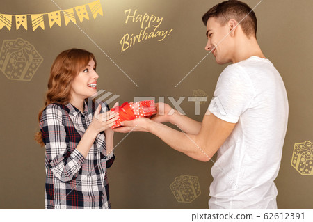 Celebration Concept. Young boyfriend giving gift to touched girlfriend standing isolated on grey close-up Celebration Concept. Young boyfriend giving gift to touched girlfriend standing isolated on grey close-up 62612391