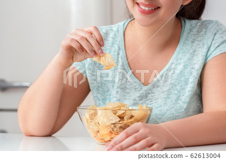 Breaking Diet. Chubby girl sitting at kitchen table eating chips smiling joyful close-up 62613004