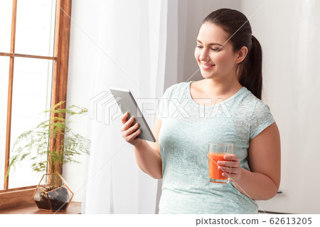 Body Care Motivation. Chubby girl standing in kitchen near window drinking detox smoothie watching video on digital tablet smiling happy Body Care Motivation. Chubby girl standing in kitchen near window drinking detox smoothie watching video on digital tablet smiling happy 62613205