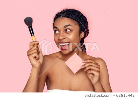 Beauty Concept. Young african woman wearing makeup isolated on pink with brush and sticker copy space explaining step smiling joyful 62613442