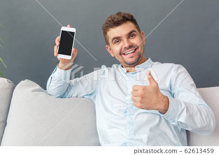 Copy Space For Product or Text. Young man studio sitting on sofa isolated on grey with smartphone showing thumb up to camera happy Copy Space For Product or Text. Young man studio sitting on sofa isolated on grey with smartphone showing thumb up to camera happy 62613459