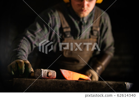 A man in protecting headphones working with hot metal knife blank 62614340
