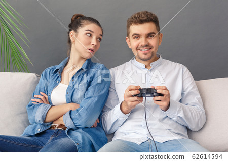 Relationnship Concept. Young couple man smiling excited playing video game sitting on sofa studio isolated on grey woman looking at him irritated Relationnship Concept. Young couple man smiling excited playing video game sitting on sofa studio isolated on grey woman looking at him irritated 62614594
