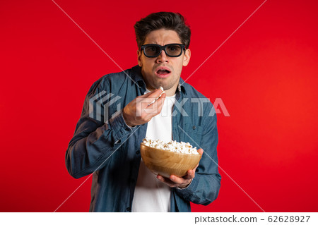 Young scared man in 3d glasses watching fascinating horror movie and eating popcorn on red studio background. Young scared man in 3d glasses watching fascinating horror movie and eating popcorn on red studio background. 62628927