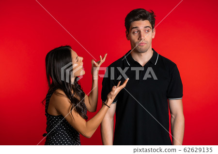 Young woman emotionally screaming at her husband or boyfriend on red background in studio. Bored man rolling his eyes. Concept of conflict, problems in relationships. Young woman emotionally screaming at her husband or boyfriend on red background in studio. Bored man rolling his eyes. Concept of conflict, problems in relationships. 62629135