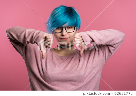 Young pretty woman with blue hair standing on pink studio background expressing discontent and showing thumb down gesture at camera. Portrait of girl with sign of dislike Young pretty woman with blue hair standing on pink studio background expressing discontent and showing thumb down gesture at camera. Portrait of girl with sign of dislike 62631773