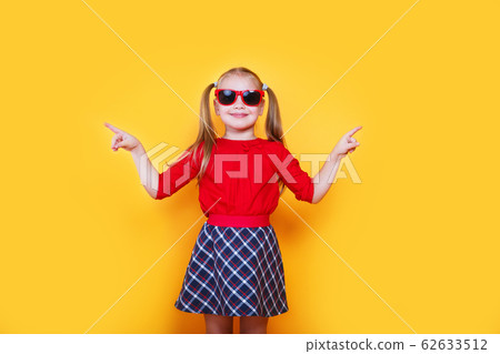 Little girl wearing sunglasses smiling and pointing to the side with thumb up. Little girl wearing sunglasses smiling and pointing to the side with thumb up. 62633512