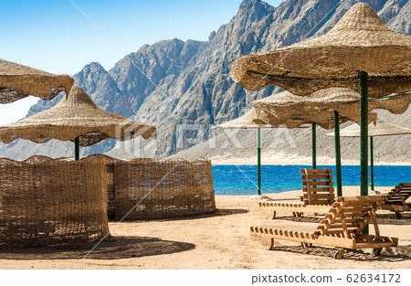 beach umbrellas and wooden lounge chairs on the sand of the beach against the backdrop of the sea and high rocky mountains 62634172