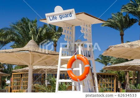 wooden lifeguard tower with orange lifebuoy on the beach against the backdrop of palm trees and reed parasols wooden lifeguard tower with orange lifebuoy on the beach against the backdrop of palm trees and reed parasols 62634533