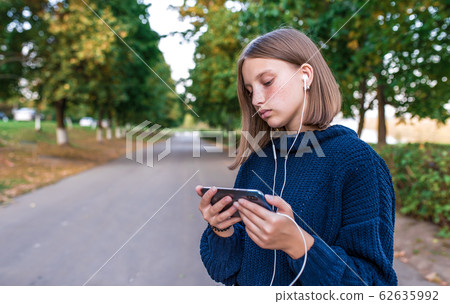 Serious teenage girl schoolgirl, summer park, holding smartphone in hand, watching video headphones. Rest in park after school college. Emotions understanding learning, video lesson. Serious teenage girl schoolgirl, summer park, holding smartphone in hand, watching video headphones. Rest in park after school college. Emotions understanding learning, video lesson. 62635992