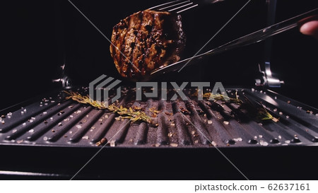 Delicious juicy meat steak cooking on grill. Aged prime rare roast grilling marble beef. Electric roaster, rosemary black pepper, salt. Kitchen tongs. 62637161