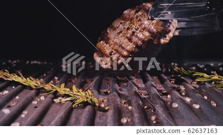 Delicious juicy meat steak cooking on grill. Aged prime rare roast grilling marble beef. Electric roaster, rosemary black pepper, salt. Kitchen tongs. Delicious juicy meat steak cooking on grill. Aged prime rare roast grilling marble beef. Electric roaster, rosemary black pepper, salt. Kitchen tongs. 62637163