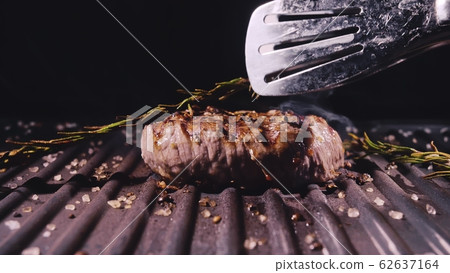 Delicious juicy meat steak cooking on grill. Aged prime rare roast grilling marble beef. Electric roaster, rosemary black pepper, salt. Kitchen tongs. Delicious juicy meat steak cooking on grill. Aged prime rare roast grilling marble beef. Electric roaster, rosemary black pepper, salt. Kitchen tongs. 62637164