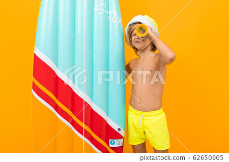 Handsome boy in swimming trunks holds a rubber mattress, smiles and gesticulates isolated on orange 62650905